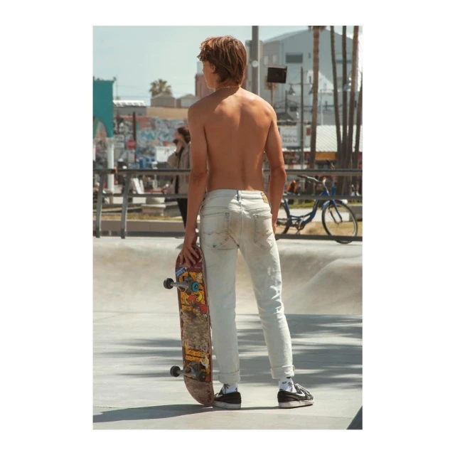 Shirtless adult male stands in a skate park holding a skateboard, wearing jeans and sneakers, with other people and palm trees in the background.