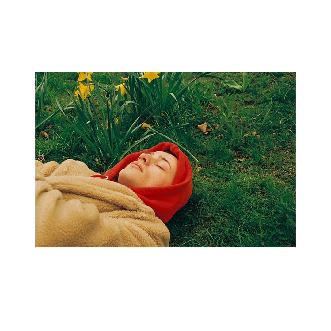 Woman in red hoodie and beige coat relaxes on green grass near yellow daffodils in natural light; outdoor springtime scene.