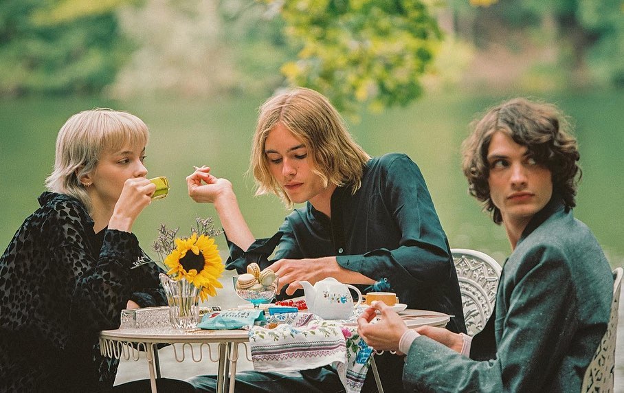 Three young adults sit outdoors around a table with a sunflower, teapot, tea cups, and snacks in front of a blurred background.