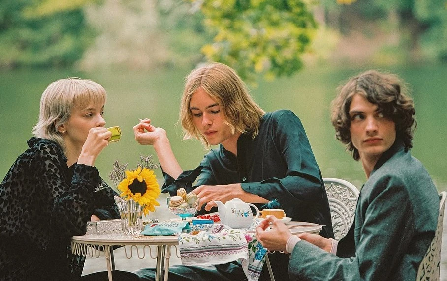 Three young adults sit outdoors around a table with a sunflower, teapot, tea cups, and snacks in front of a blurred background.