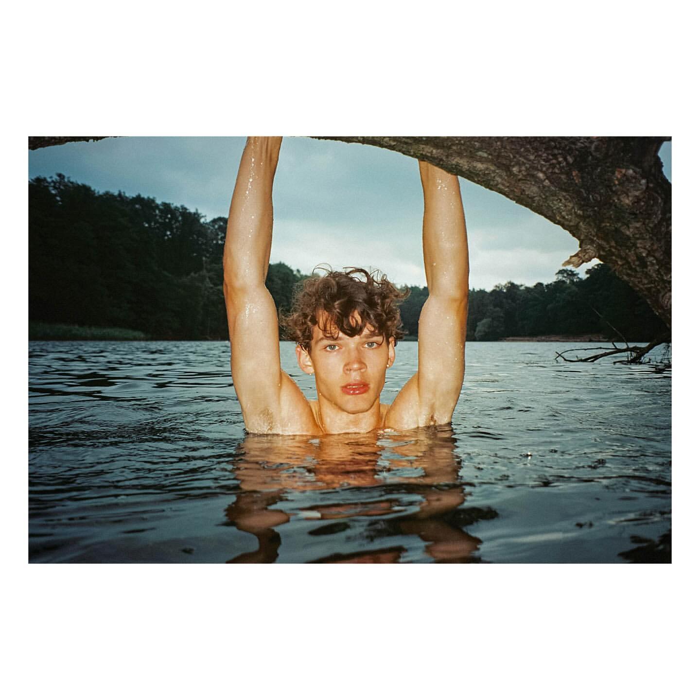 Young man with curly hair submerged in a lake with his arms raised, framed by a tree branch overhead.