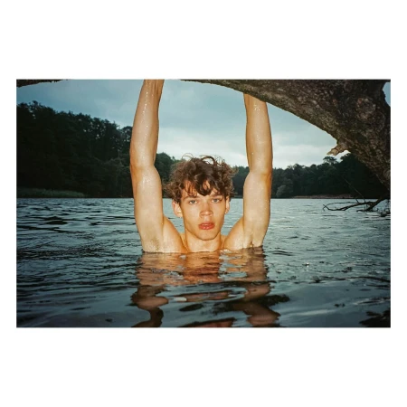 Young man with curly hair submerged in a lake with his arms raised, framed by a tree branch overhead.