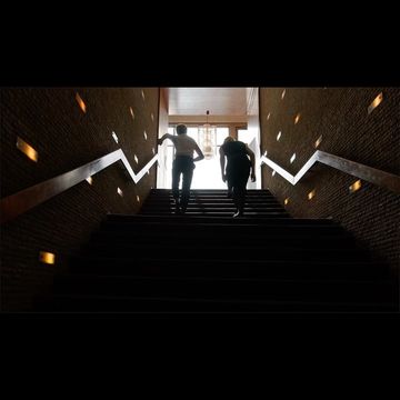 Two silhouetted figures walking up a wide dark indoor staircase with lit wall sconces by Niv Shank.