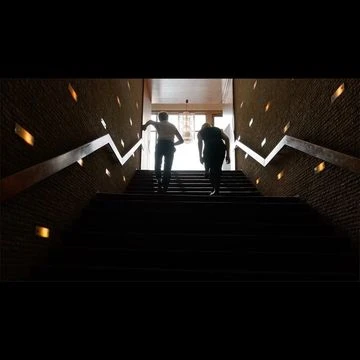 Two silhouetted figures walking up a wide dark indoor staircase with lit wall sconces by Niv Shank.