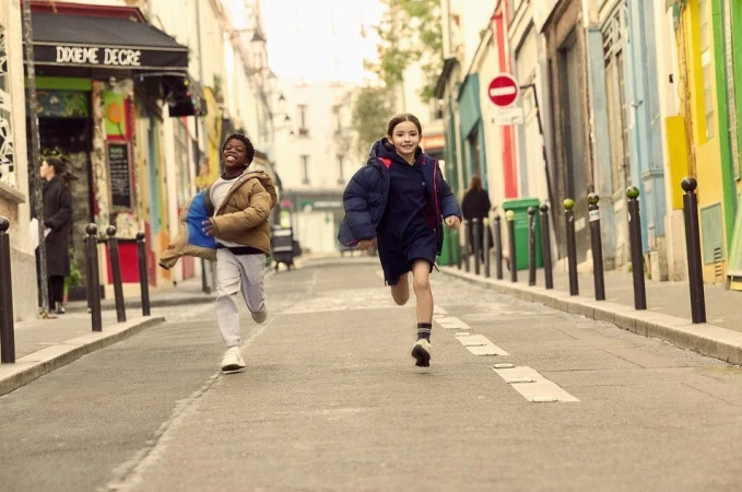 Two children, one African American and one White, run down a colorful street in puffy jackets, capturing a lively and youthful energy.