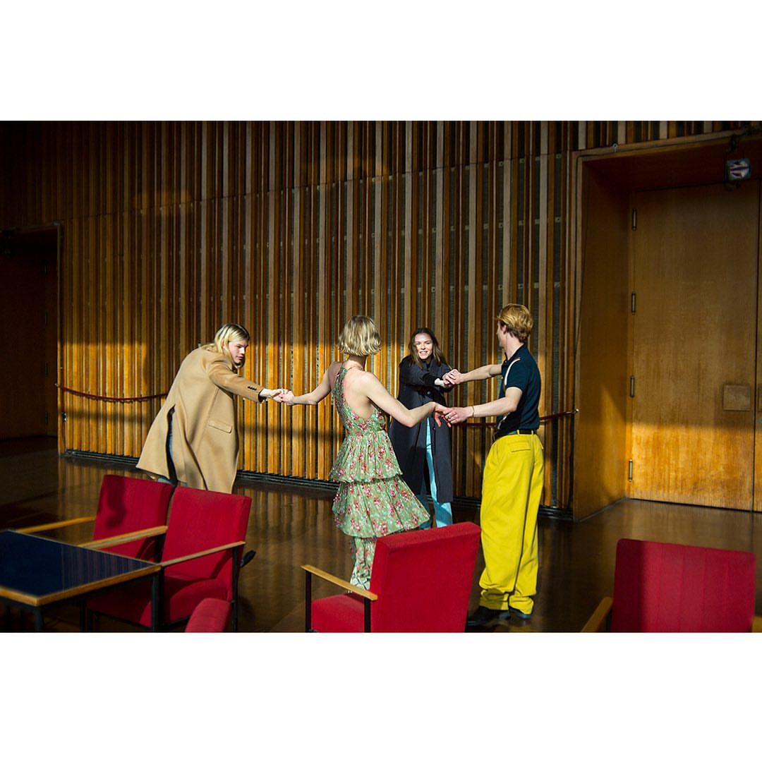 Four people in a bright indoor space with wooden walls, red chairs, and large doorway. Two women hold hands in center.