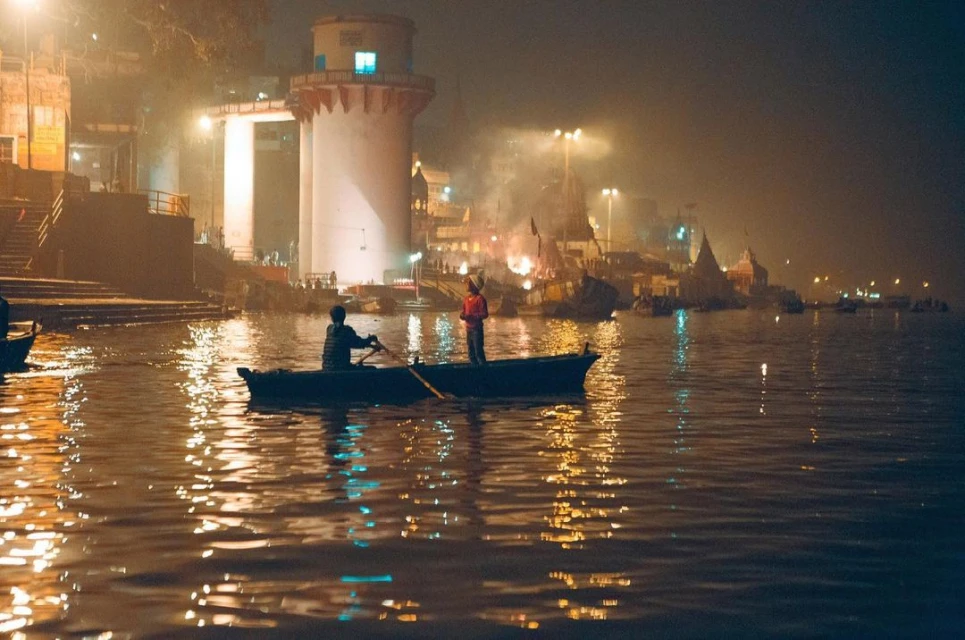 Two people in a rowboat at night on a river with lights reflecting on the water, near buildings on the riverbank under a smoky sky.