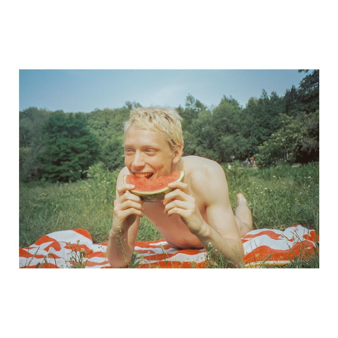 Blond-haired man lying on a striped blanket in a grassy field, smiling while holding a slice of watermelon up to his mouth.