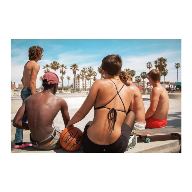 Group of young people gather on a concrete surface near a skate park on a bright, sunny day with palm trees and buildings.