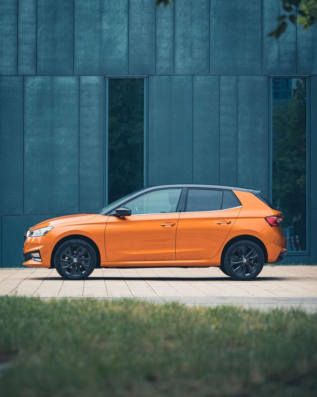 Orange Skoda Fabia hatchback car parked outdoors against a modern building with green grass in the foreground on a sunny day.