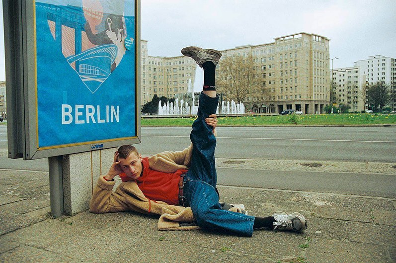 Man lounges on sidewalk next to Berlin travel advertisement. One leg is raised high in the air. A fountain and buildings are in the background.