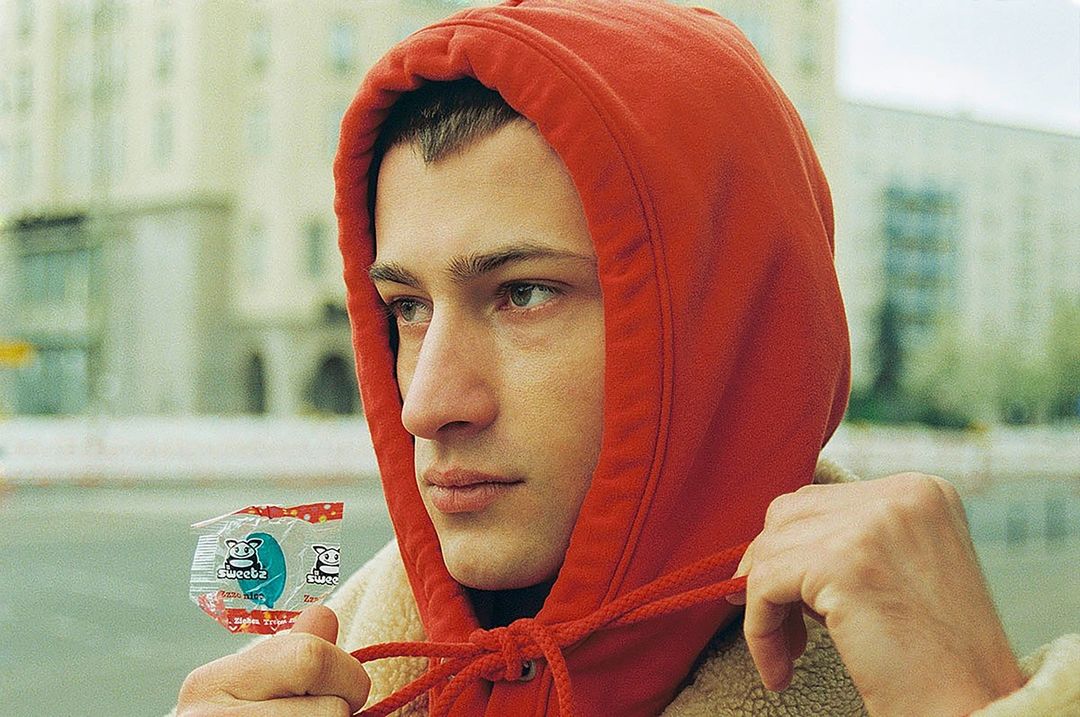 Close-up of a young adult man wearing a red hoodie and holding a candy wrapper outdoors with buildings in the background.