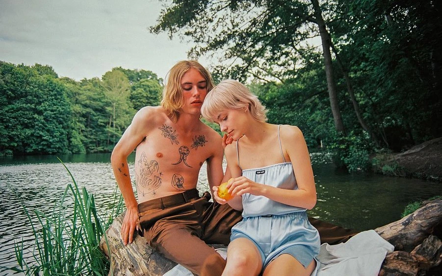 An outdoor fashion editorial featuring a blonde woman and a man with visible tattoos sitting on a log near a lake and forest.