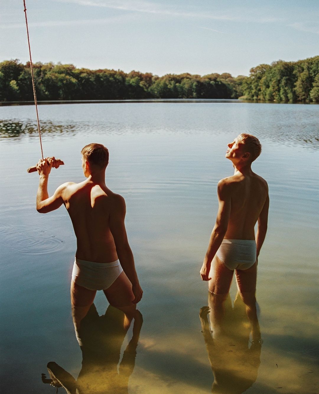 Two adult men stand in lake with trees in background. One man holds a rope, the other looks up. Natural light, outdoor setting, summer day.