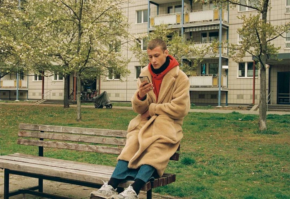 An adult male sits on a park bench looking at his smartphone, wearing a red hoodie, jeans, and a long tan coat in daytime.