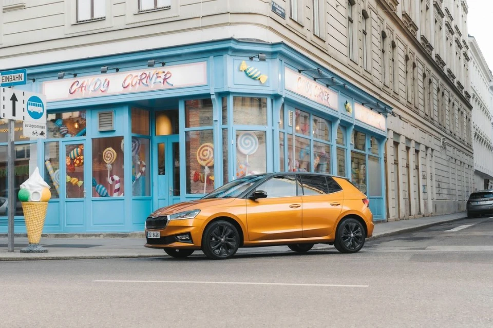 Orange Skoda Fabia parked in front of Candy Corner store in European city setting with street sign and cone statue visible.