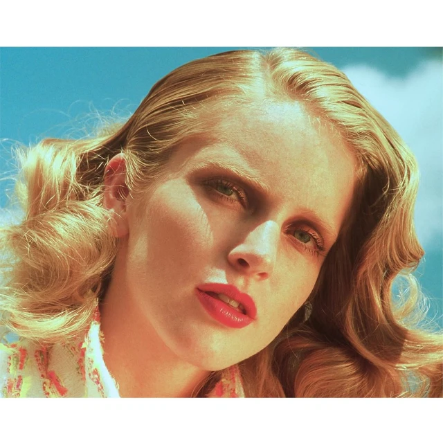 Close-up portrait of a blond adult woman with red lipstick, styled hair, wearing a patterned top against a light blue sky.