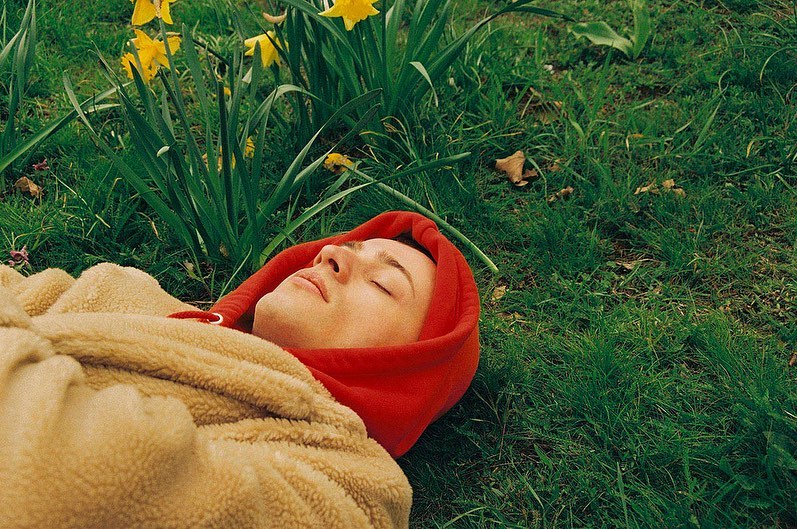 Adult laying in grass with yellow daffodils wearing a red hooded sweatshirt and a tan sherpa-like jacket, eyes closed, outdoor setting.