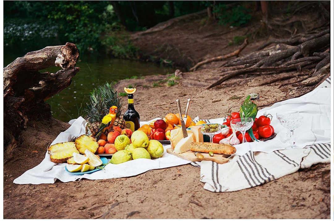 Outdoor picnic still life with fresh fruit, cheese, bread, and wine arranged on a white cloth, set on sandy ground, with a leafy background.