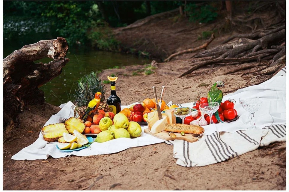 Outdoor picnic still life with fresh fruit, cheese, bread, and wine arranged on a white cloth, set on sandy ground, with a leafy background.