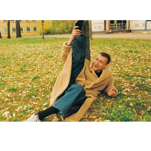 Young man in a beige coat lying on grassy ground covered in leaves, extending his leg in the air, smiling with trees and building behind.