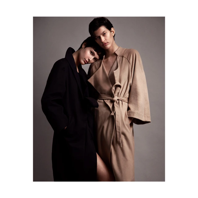 Two models in a photo studio. One model rests head on the other model's shoulder. Both wear fashionable robe coats, one black and one tan.
