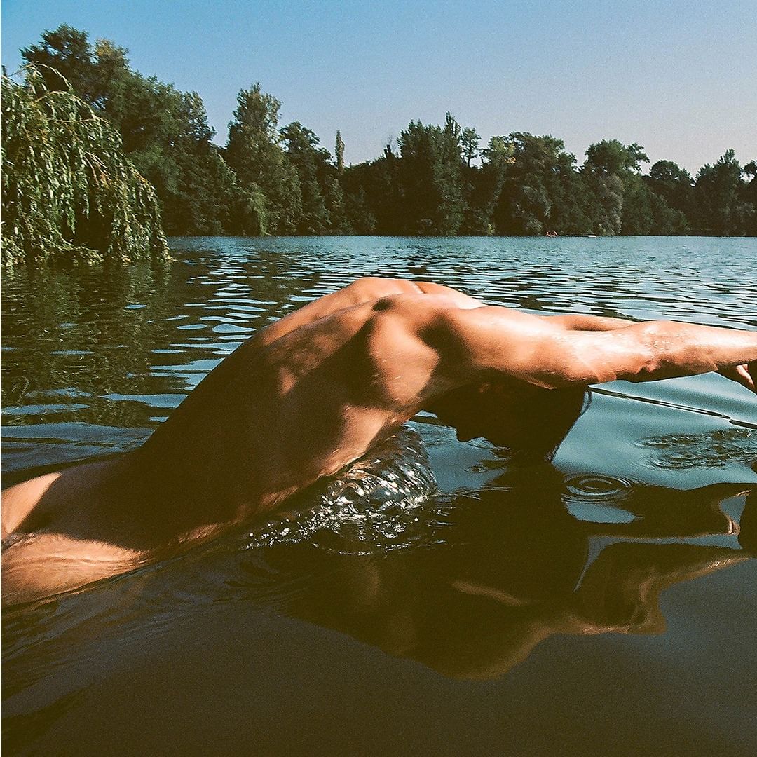 Torso and arm of an adult male in a lake, swimming with a tree-lined shoreline, and a clear sky on a sunny day.