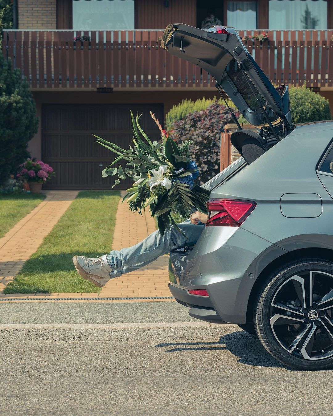 Person with legs sticking out the back of a gray hatchback car with a bouquet of flowers, in front of a brown house and green lawn.