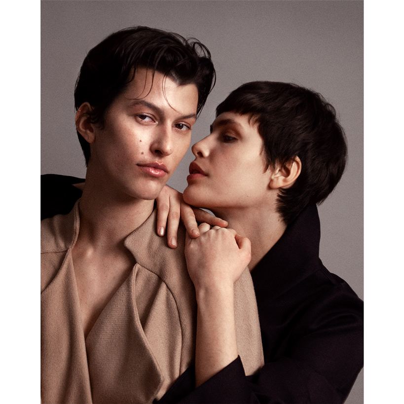 Fashionable couple poses closely in a studio setting, with one person in a tan coat and the other in a dark coat, showing an intimate moment.