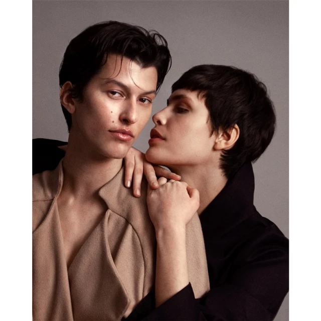 Fashionable couple poses closely in a studio setting, with one person in a tan coat and the other in a dark coat, showing an intimate moment.