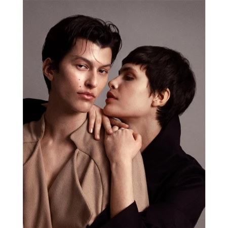 Fashionable couple poses closely in a studio setting, with one person in a tan coat and the other in a dark coat, showing an intimate moment.