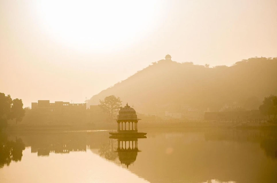 Scenic view of a domed pavilion reflected in a tranquil lake at sunset, with a backdrop of hills and a historic building.