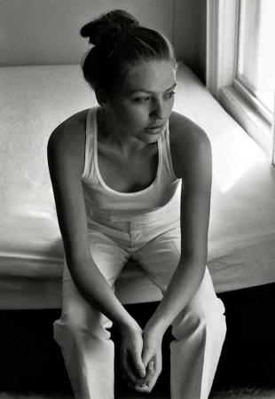Monochrome portrait of a woman in a white tank top and trousers seated on a bed near a window.