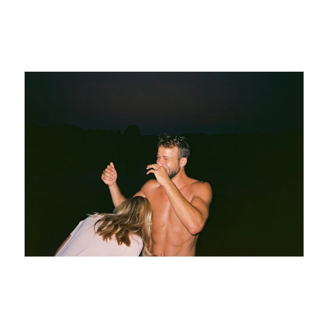Shirtless man laughing outdoors with a woman in a light pink shirt, low light, against a dark background in a medium outdoor shot.