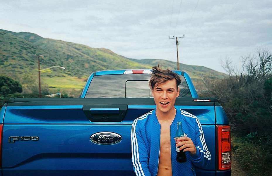 Young man smiling, holding a Coca-Cola bottle in front of a blue Ford F-150 truck against a background of green hills.