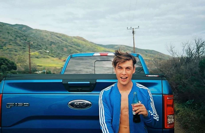 Young man smiling, holding a Coca-Cola bottle in front of a blue Ford F-150 truck against a background of green hills.