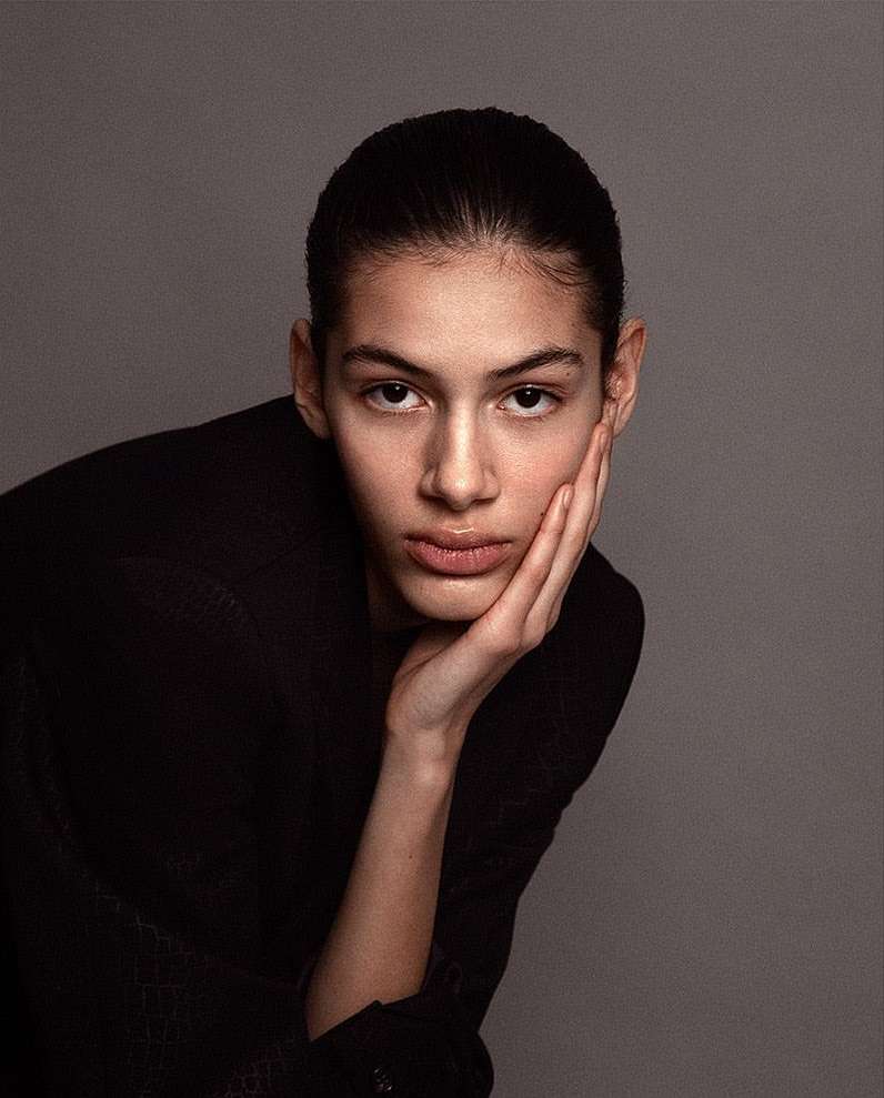 Close-up portrait of a model with dark hair wearing a black jacket, resting chin on hand against a neutral grey background.
