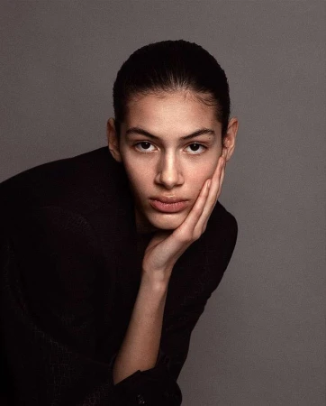 Close-up portrait of a model with dark hair wearing a black jacket, resting chin on hand against a neutral grey background.