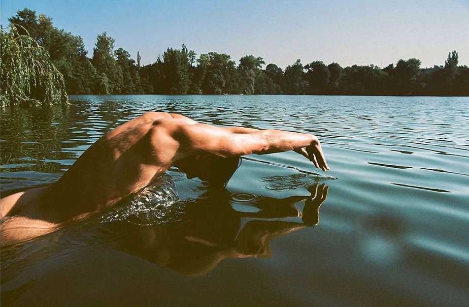 Person swimming in a lake with trees visible in the background and blue sky above, creating a serene outdoor scene.