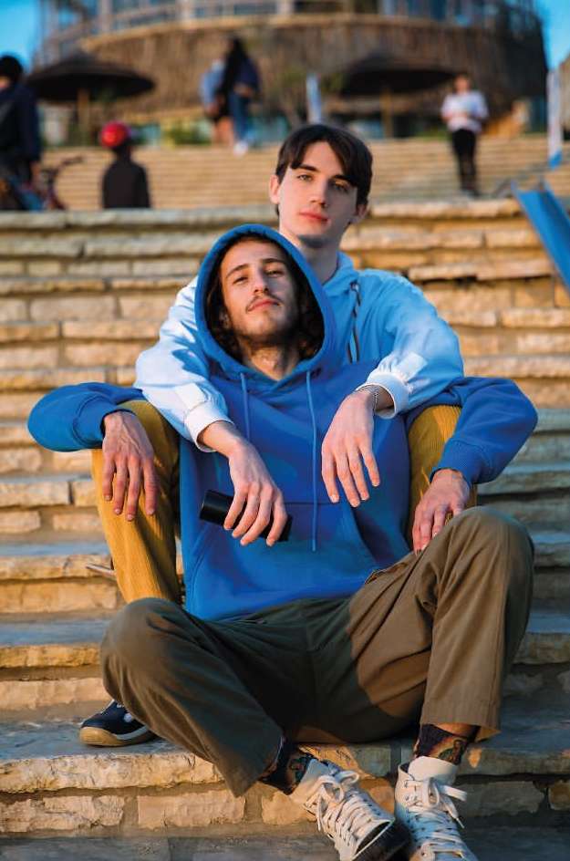 Two young men pose outdoors on concrete steps, one seated in a blue hoodie, the other standing behind with a light blue hoodie.