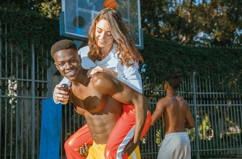 Young man carries a smiling woman on his back near a basketball hoop in an outdoor setting with another person in the background.
