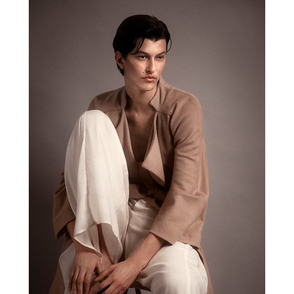 Model with dark hair wearing a tan jacket and white trousers seated against a grey backdrop