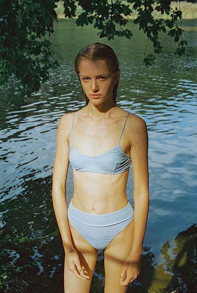 Young woman in a swimsuit standing in water near the shoreline with dappled sunlight and green foliage in a natural setting.