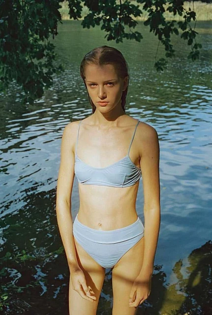 Young woman in a swimsuit standing in water near the shoreline with dappled sunlight and green foliage in a natural setting.