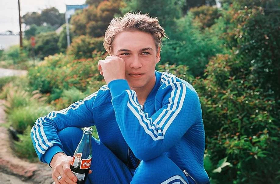 Young man in a blue tracksuit sitting and holding a Coca-Cola bottle, posing outdoors among trees and foliage in natural daylight.