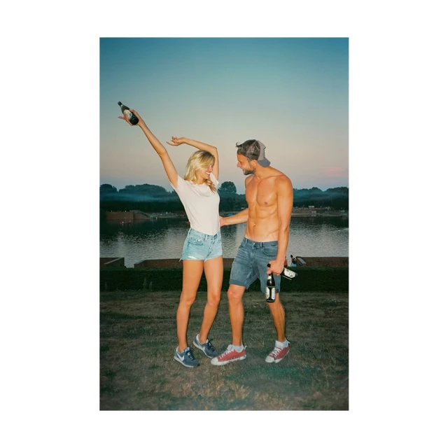 A joyful couple is enjoying a sunny day outdoors, holding bottled drinks near a lake, showcasing a casual and celebratory moment.