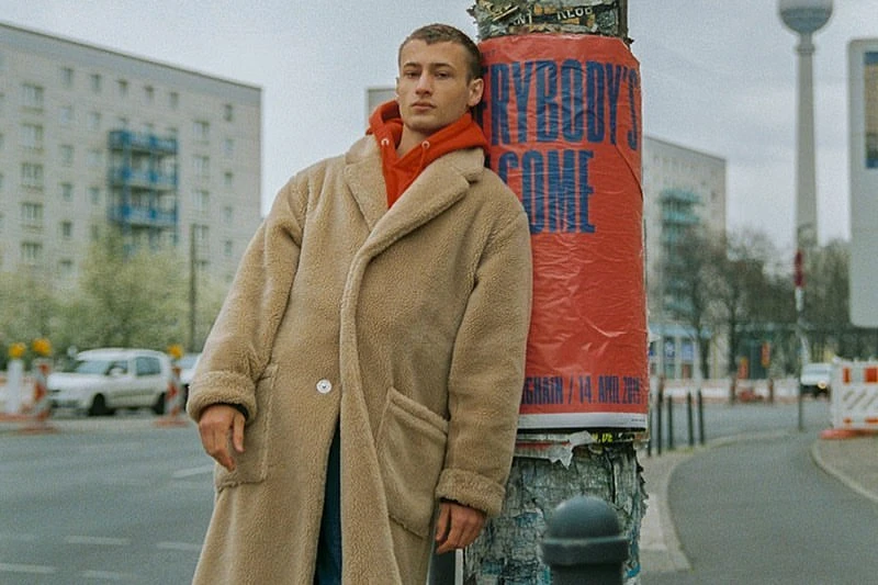 A young adult man wearing a long beige coat and a red hoodie leans against a telephone pole in an urban setting, creating a street-style look.