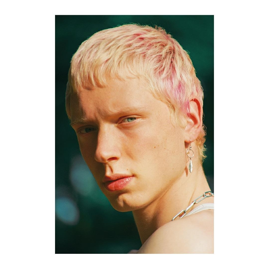 Portrait of a young adult with light skin, dyed blonde and pink hair, wearing a silver necklace and a dangling earring, against green background.