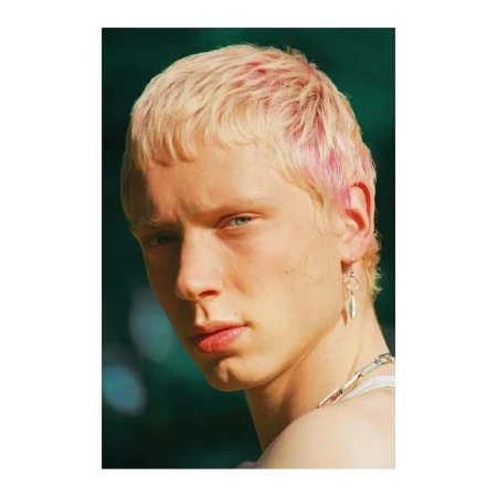 Portrait of a young adult with light skin, dyed blonde and pink hair, wearing a silver necklace and a dangling earring, against green background.