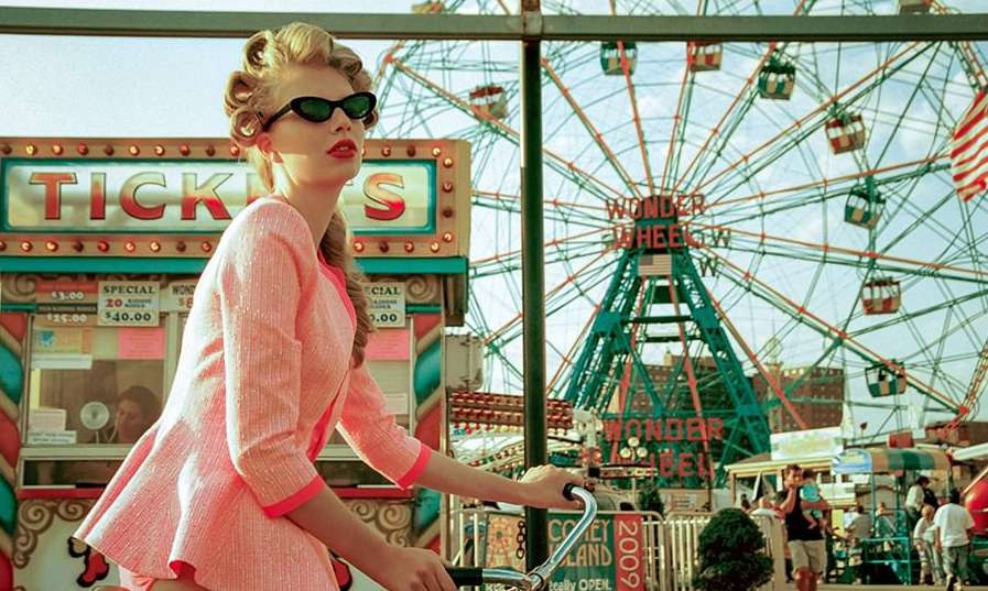 Blonde woman in pink dress and sunglasses riding a bicycle in front of a colorful Ferris wheel and ticket booth at Coney Island.